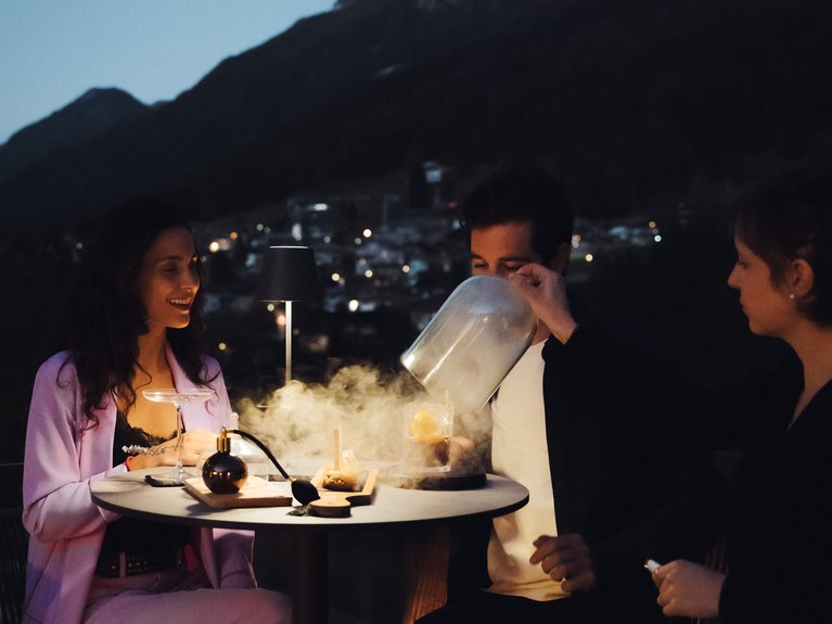 Hotel Latemar mit Dolomitenblick Personen genießen dampfende Cocktails auf einer nächtlichen Terrasse in den Bergen