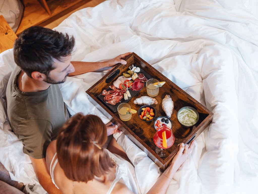 Hotel Latemar mit Dolomitenblick Ein Paar mit Frühstück im Bett mit Wurst, Croissant, Obst und Getränken