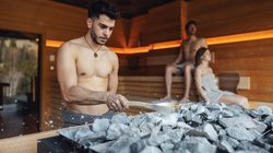 Hotel Latemar mit Dolomitenblick Mann gießt Wasser auf heiße Steine in der Sauna mit zwei Personen im Hintergrund
