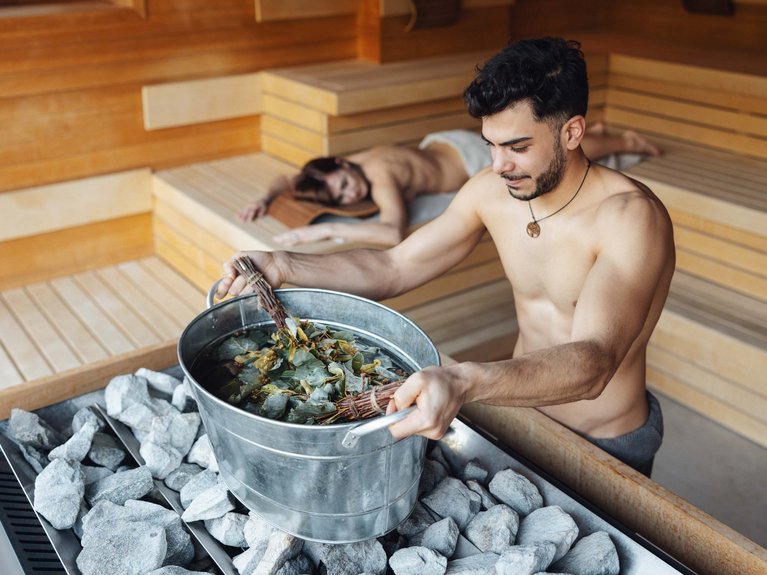Hotel Latemar mit Dolomitenblick Mann mit Eimer voller Kräuter in der Sauna, Frau entspannt im Hintergrund