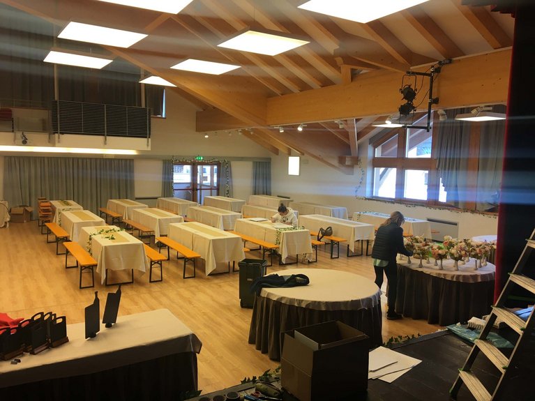 On the webcam Latemar is lovely, in real life it’s a dream! Room with tables and benches set for event, two people arranging decorations