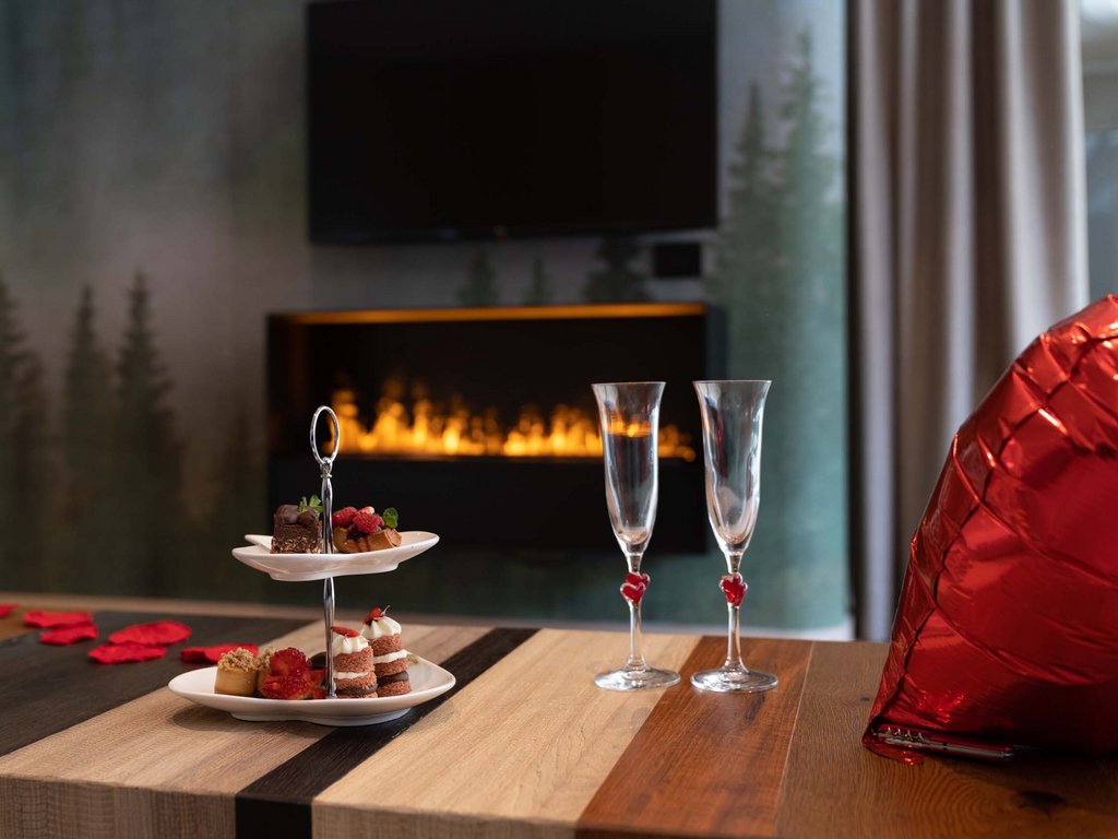 Val di Fassa: wandern, biken, erleben Tisch mit Kuchen, zwei Gläsern und rotem Herzballon vor einem brennenden Kamin