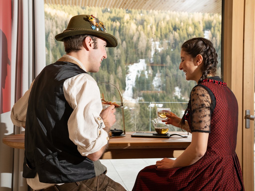 Sulla pista ciclabile, da Moena alla vostra oasi romantica Coppia in abiti tradizionali che beve tè vicino a una finestra con vista su foresta e neve
