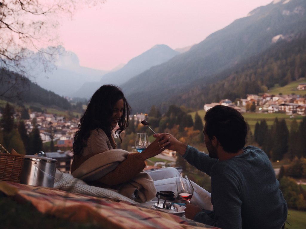 Sulla pista ciclabile, da Moena alla vostra oasi romantica Coppia che fa picnic in montagna al tramonto con vista sul villaggio