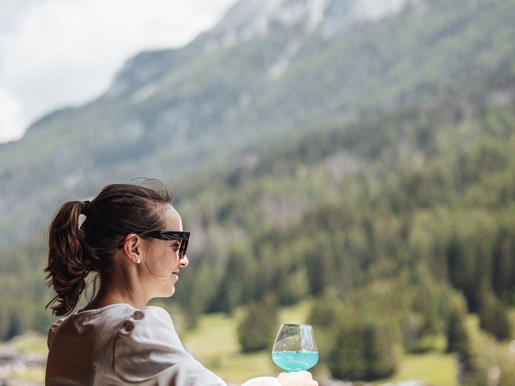 Barrierefreies Hotel im Val di Fassa: das Latemar Frau mit Sonnenbrille hält ein blaues Glas auf einem Balkon mit Bergblick