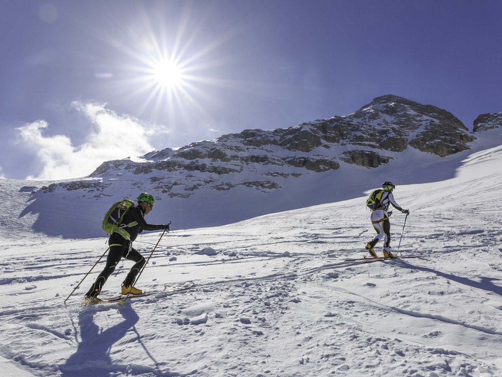 Skigebiet Fassatal Pistenplan Zwei Skifahrer mit Rucksäcken steigen bei Sonnenschein einen verschneiten Berg hinauf
