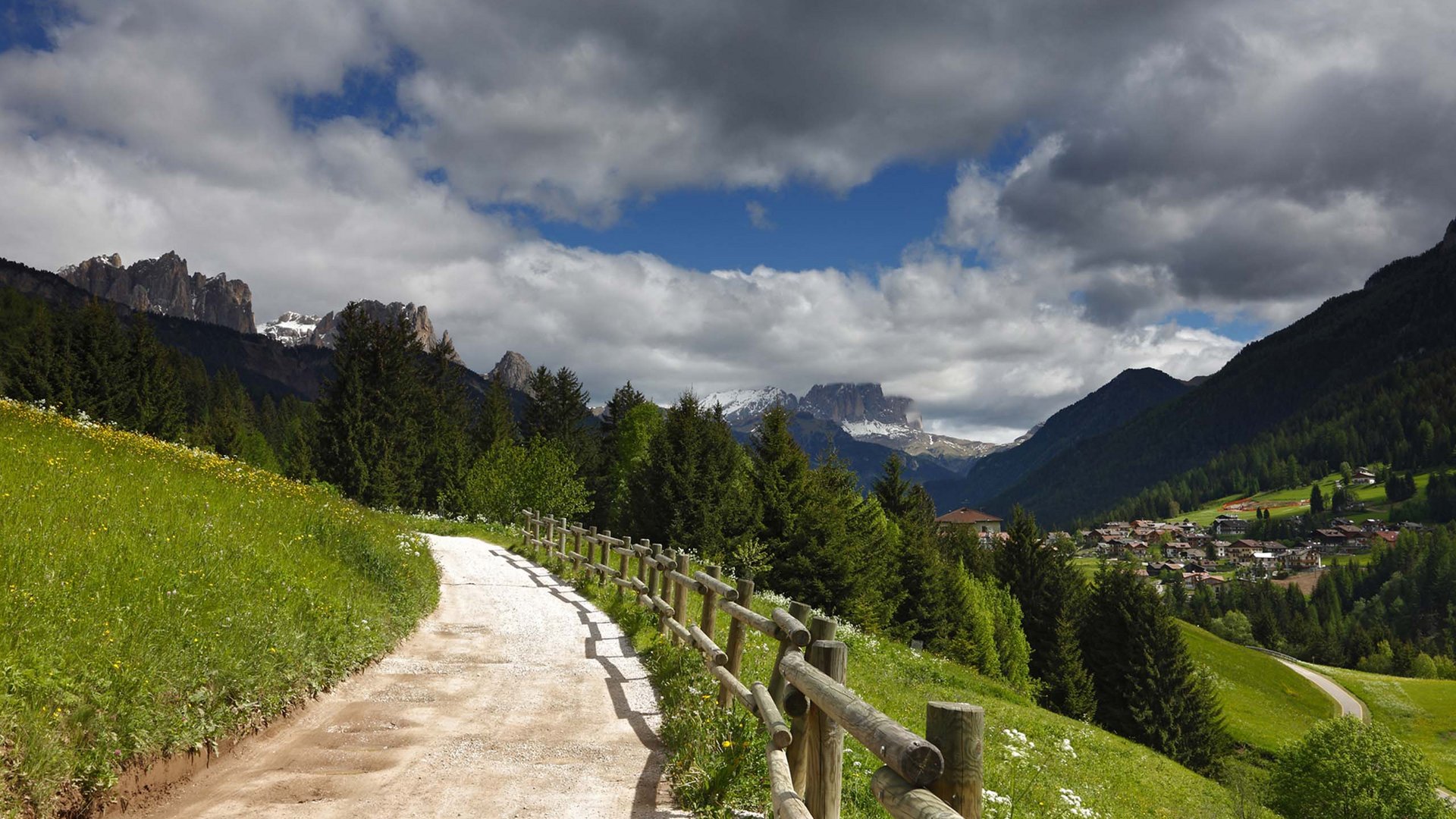 Noleggio bici a Soraga: un must in autunno all’Hotel Latemar Sentiero di campagna con recinzione in legno tra colline verdi e montagne con villaggio