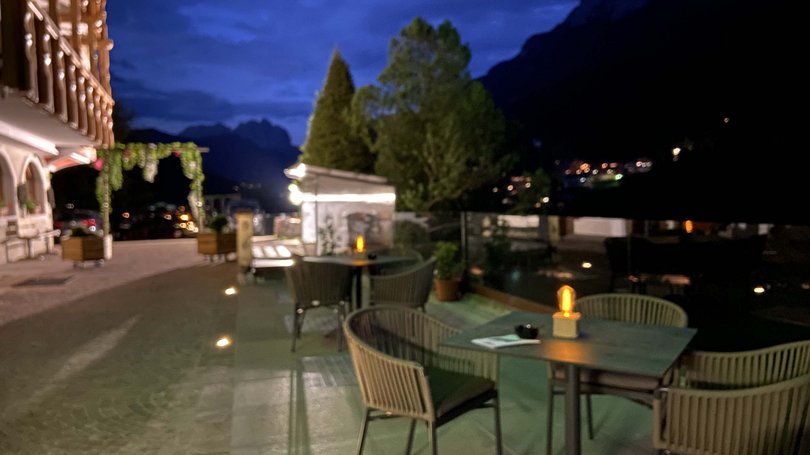 Moena: Unterkunft für Ihren Traumurlaub. Hotel Latemar! Abendliche Terrasse mit Tischen und Stühlen neben Gebäude und Bäumen beleuchtet