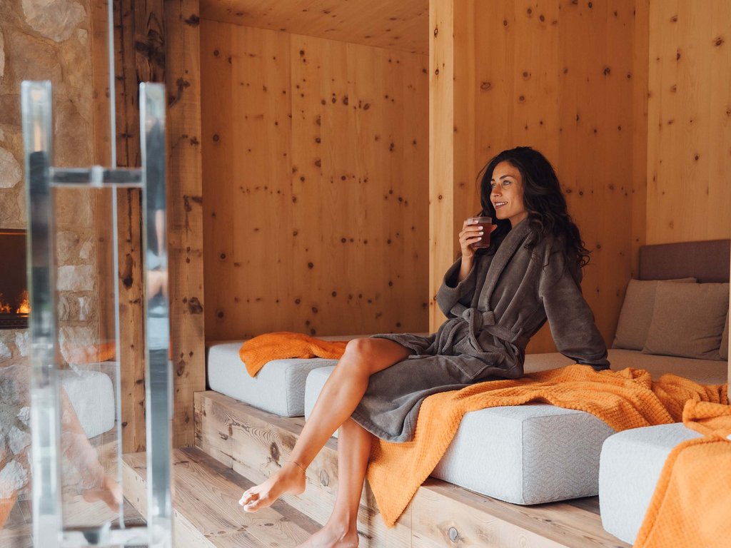 Trentino: Hotel am See mit Pool und Spa Frau im grauen Bademantel trinkt Getränk, sitzt in holzvertäfeltem Spa-Bereich