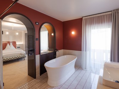 Barrierefreies Hotel im Val di Fassa: das Latemar Modernes Badezimmer mit weißer Badewanne, Spiegeln und sichtbarem Schlafzimmer