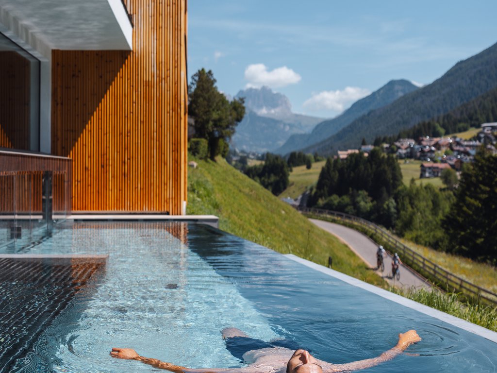 A Soraga il meteo è sempre ottimo per un Day Spa! Uomo che galleggia in piscina a sfioro con vista sulle montagne verdi e cielo limpido