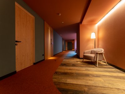 Barrierefreies Hotel im Val di Fassa: das Latemar Hotelflur mit Holzfußboden, rotem Teppich und beleuchtetem Sessel