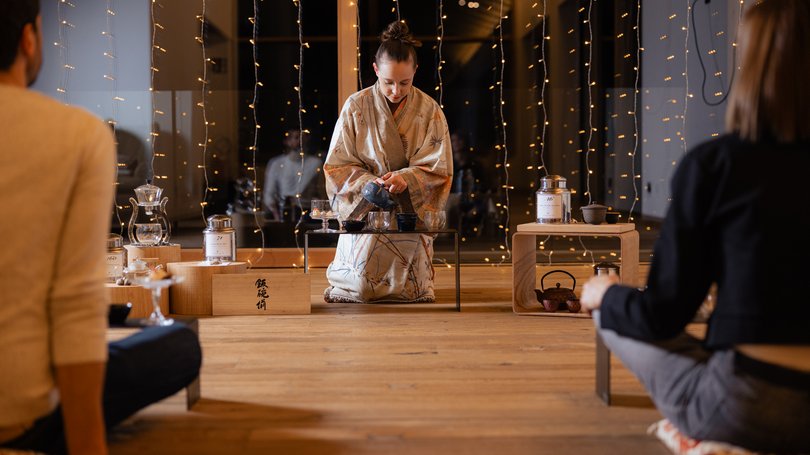 Val di Fassa: wandern, biken, erleben Frau im Kimono bereitet japanische Teezeremonie vor, zwei Personen sitzen davor.