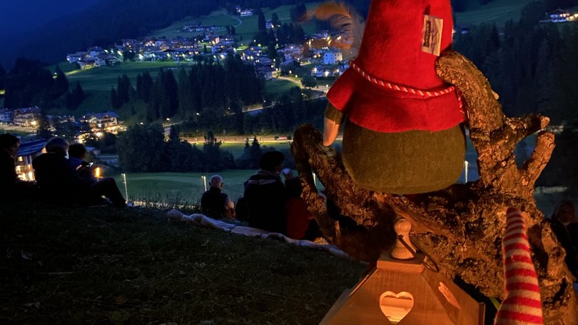 Val di Fassa: wandern, biken, erleben Leuchtende Laterne und Figuren mit roten Hüten auf Hügel bei Nacht mit Dorflichtern