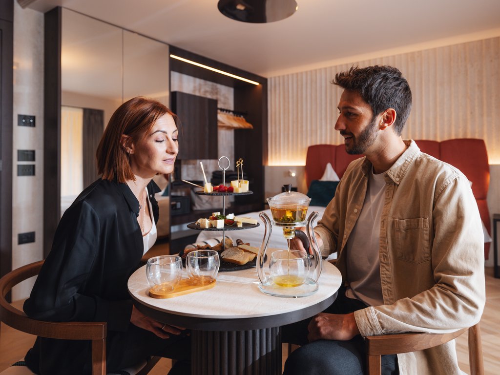 Sulla pista ciclabile, da Moena alla vostra oasi romantica Coppia che gusta tè e snack su tavolo rotondo in una stanza moderna.