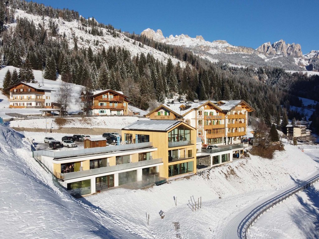 Val di Fassa: dove dormire e vivere vacanze sostenibili Hotel immerso nella neve con montagne e alberi sullo sfondo.