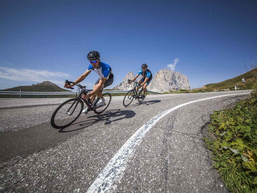 Lago di Soraga and summer activities near Hotel Latemar Two cyclists descending a curved mountain road with peaks in the background