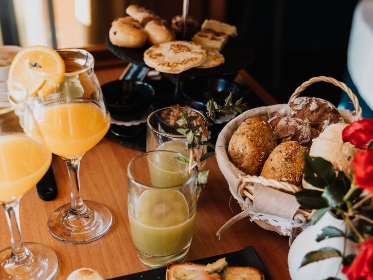 I nostri fornitori di fiducia: solo prodotti di qualità! Colazione con toast, succhi, pane, affettati e fiori rossi su tavolo di legno