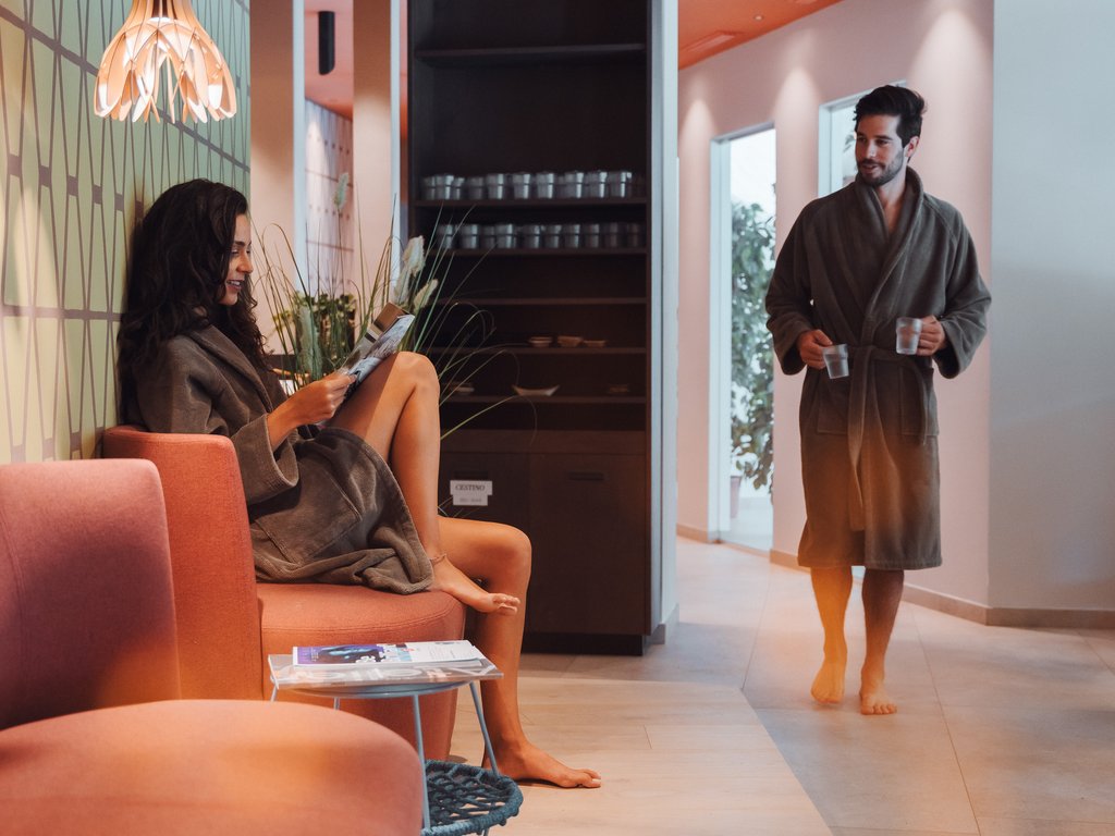 A Soraga il meteo è sempre ottimo per un Day Spa! Donna seduta su divano rosa che legge, uomo in accappatoio con bevande in mano
