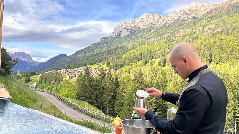 Escursioni da Moena e molto altro: il nostro programma Barista prepara un drink vicino a una piscina con vista sulle montagne verdi