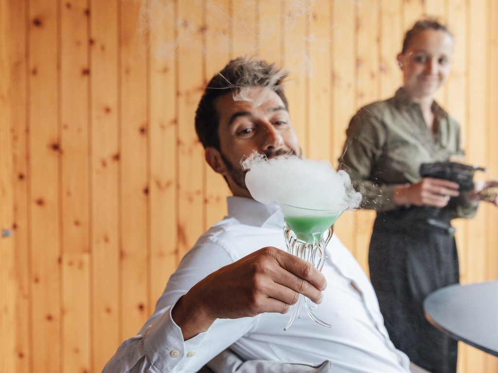 Sulla pista ciclabile, da Moena alla vostra oasi romantica Uomo sorseggia cocktail verde fumante seduto, donna sullo sfondo con utensili