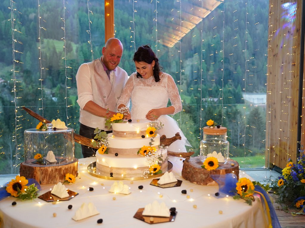 Val di Fassa: wandern, biken, erleben Glückliches Brautpaar schneidet Hochzeitstorte mit Sonnenblumen und Lichterketten