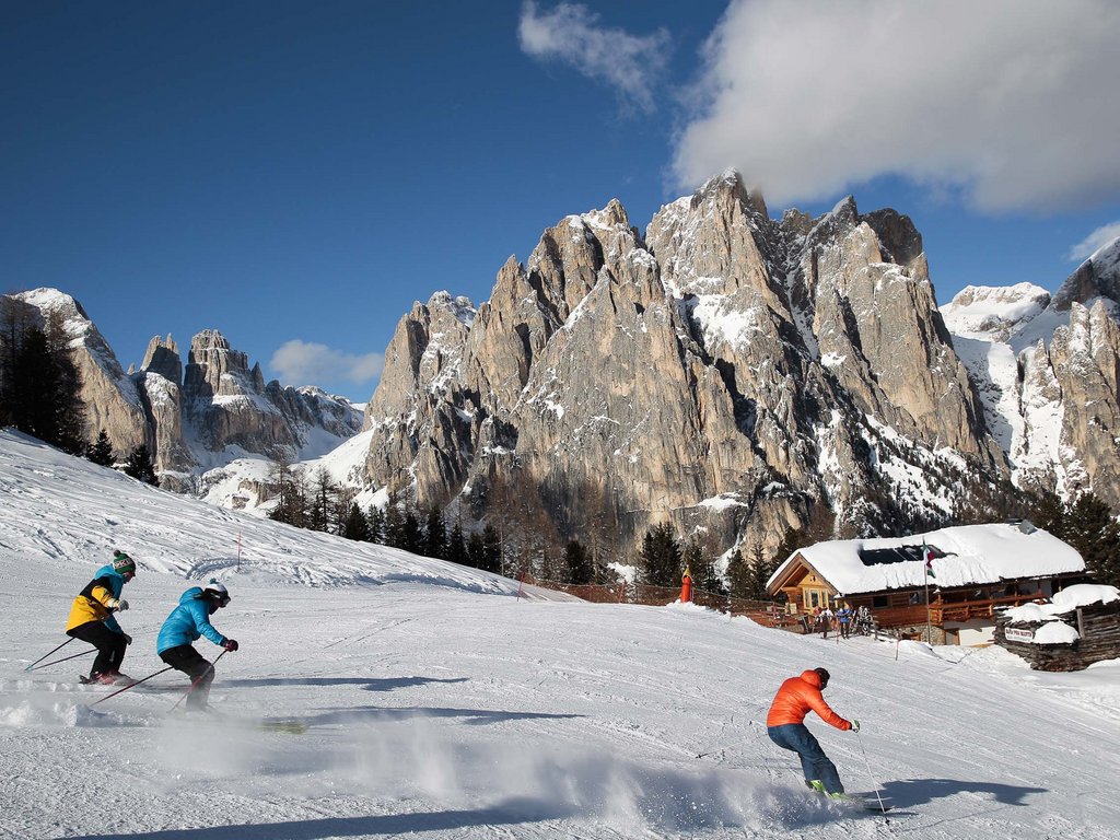 Skigebiet Fassatal Pistenplan Skifahrer fahren eine schneebedeckte Piste mit felsigen Bergen im Hintergrund hinab