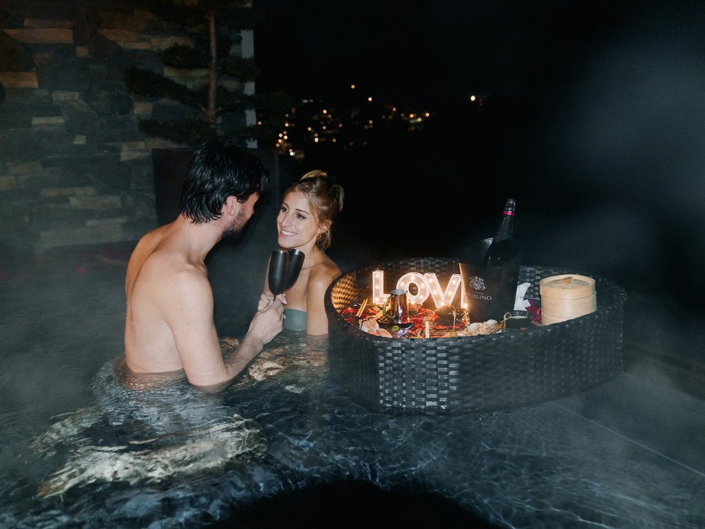 Urlaub im Fassatal: Erholung pur Paar in nächtlichem Whirlpool mit Gläsern und schwimmendem romantischem Dinner