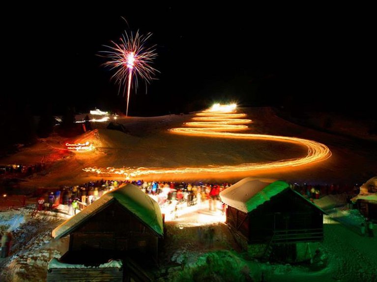 Dalla webcam il Latemar è bello, ma dal vivo è un sogno! Folla di persone e fuochi d'artificio su pista innevata con scia luminosa a zig zag di notte