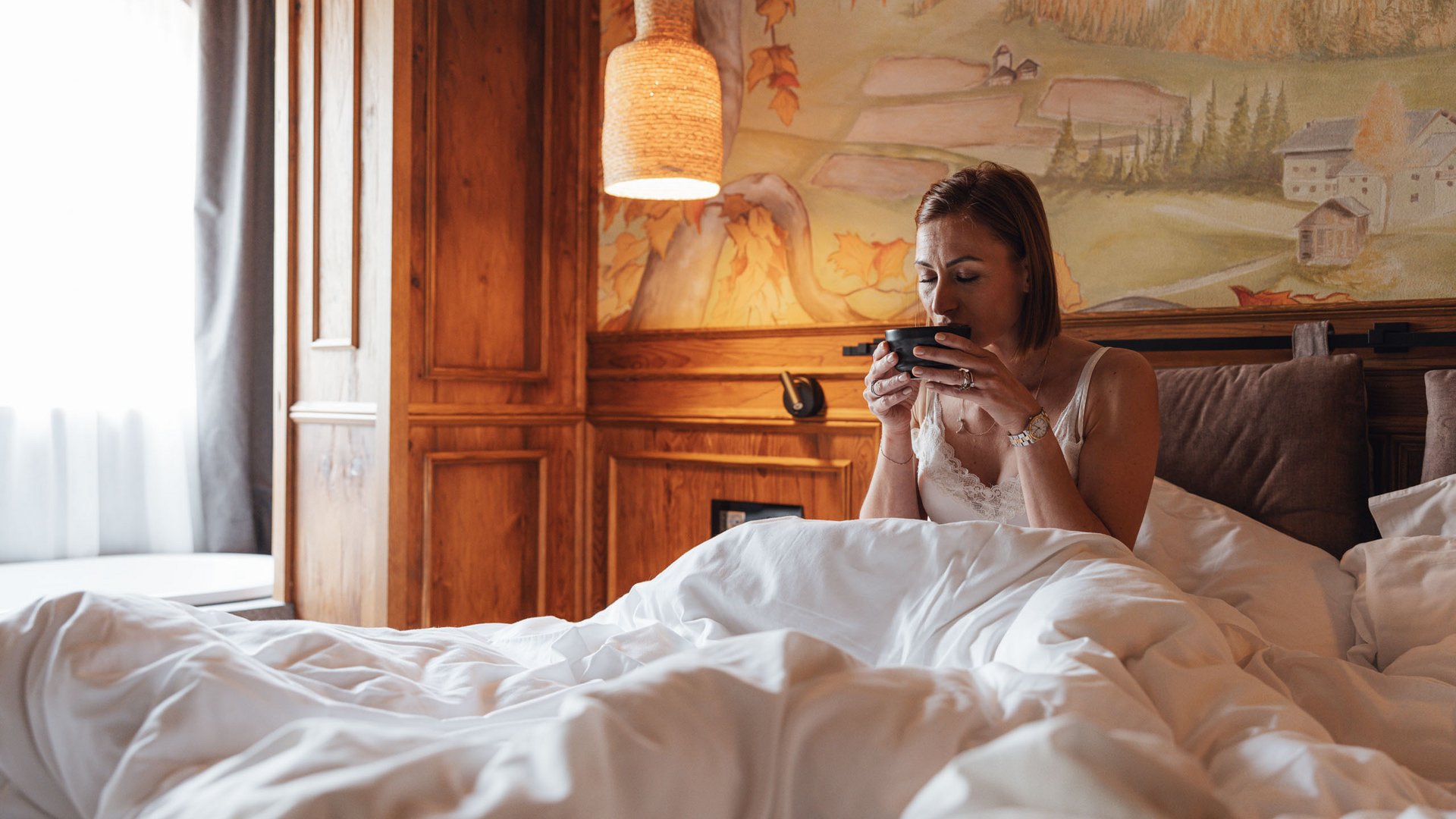 Ihr Hotel in Soraga di Fassa: Hotel Latemar Frau sitzt im gemütlichen Bett und trinkt aus einer schwarzen Tasse im rustikalen Zimmer