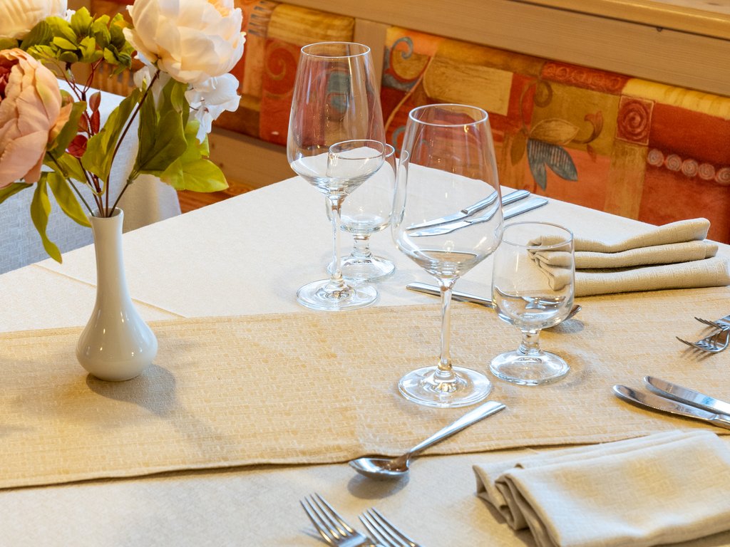 Passeggiate in Val di Fassa in inverno e pietanze deliziose Tavolo apparecchiato con bicchieri, posate e un vaso con fiori in interno rustico