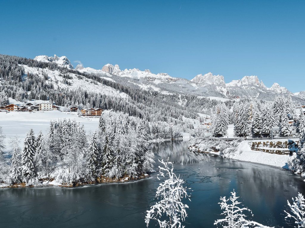 Val di Fassa: l’Hotel Latemar si trova qui Paesaggio innevato con alberi, fiume e montagne sullo sfondo