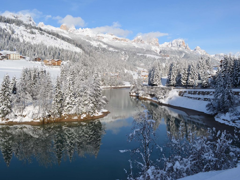 Hotel in Val di Fassa: your Hotel Latemar Winter landscape with river, snow-covered pine trees, and mountain cabins