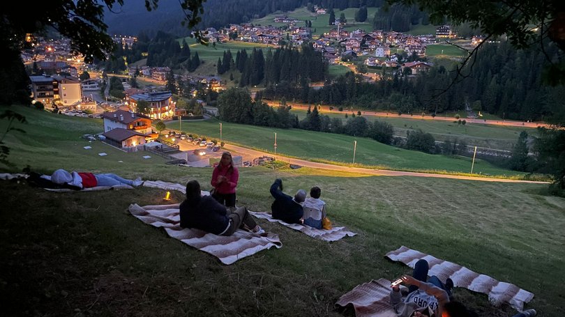 Val di Fassa: wandern, biken, erleben Menschen liegen auf Decken am Hang mit Blick auf ein abendlich beleuchtetes Dorf