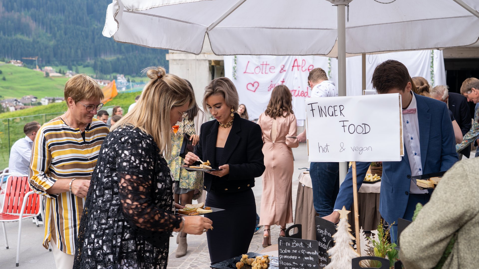Dalla webcam il Latemar è bello, ma dal vivo è un sogno! Persone che si servono cibo vegan e caldo sotto un ombrellone bianco