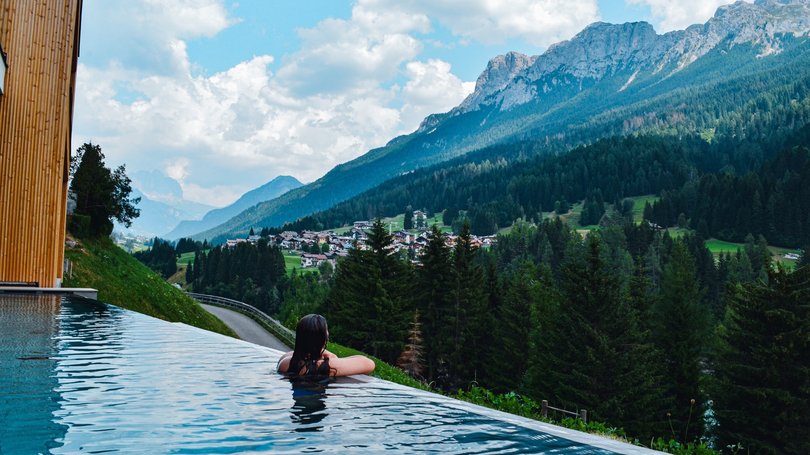 A Soraga il meteo è sempre ottimo per un Day Spa! Persona in piscina a sfioro con vista panoramica sulle montagne e villaggio