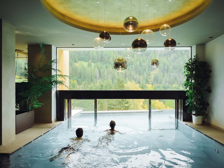 Hotel Latemar mit Dolomitenblick Innenpool mit zwei Personen und Bergblick durch ein großes Fenster