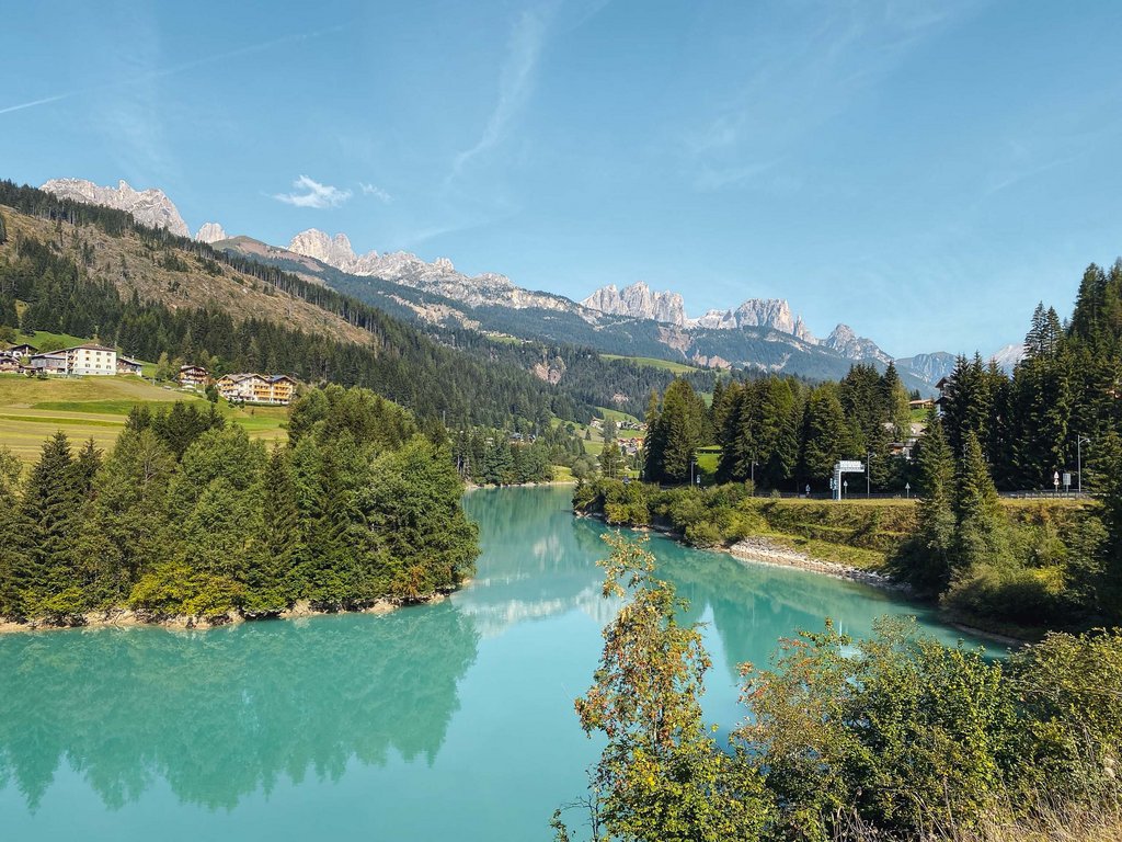 Hotel in Val di Fassa: your Hotel Latemar Blue river with forest and mountains in the background under clear sky