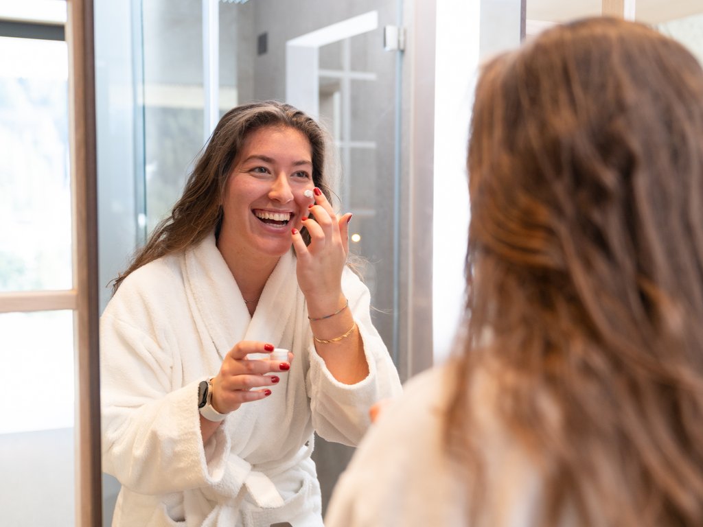 Soraga: dal lago alla piscina del Latemar Donna sorridente in accappatoio applica crema viso davanti allo specchio in bagno