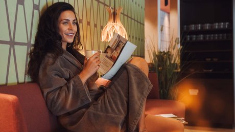 Meteo, Moena, Soraga, Latemar: tutto nella nostra gallery Donna in accappatoio rilassata su divano, legge un libro e tiene una tazza