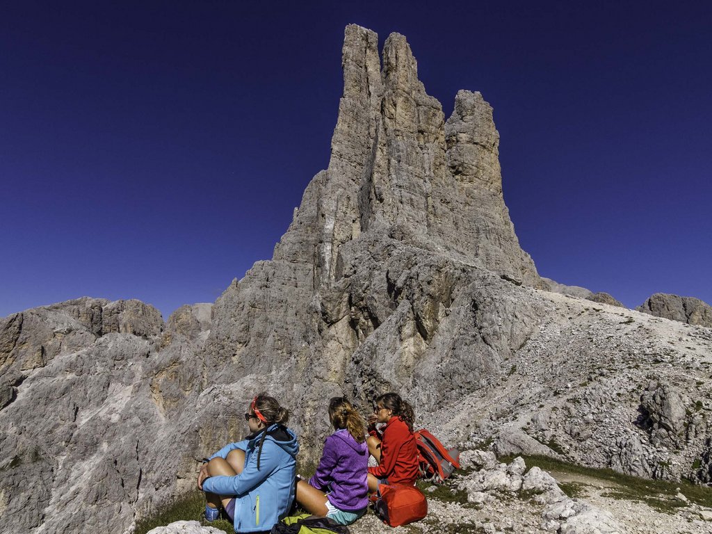Lago di Soraga and summer activities near Hotel Latemar Three hikers sitting on rocks near a rock formation under a clear sky