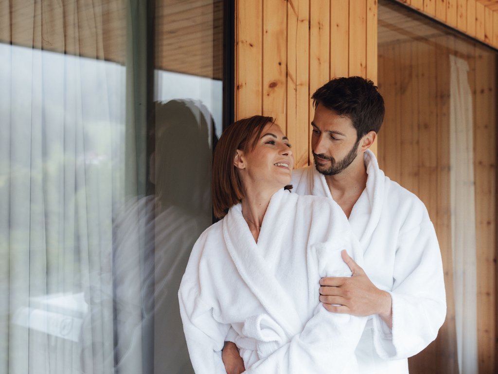 Moena: Unterkunft für Ihren Traumurlaub. Hotel Latemar! Paar in weißen Bademänteln umarmt sich neben einem Fenster in einem Holzhaus