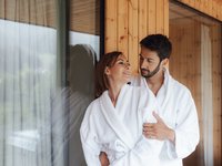 Soraga di Fassa: Wetter Paar in weißen Bademänteln umarmt sich neben einem Fenster in einem Holzhaus
