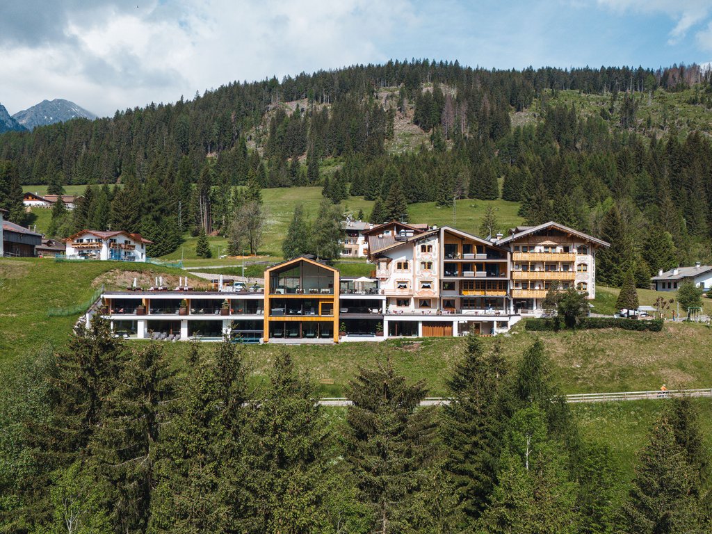 Soraga di Fassa: Urlaub in und mit der Natur Modernes Hotel in den Bergen umgeben von Bäumen und grünen Hügeln unter bewölktem Himmel