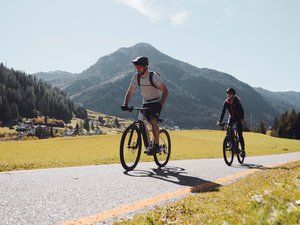 Hotel in Val di Fassa: your Hotel Latemar Two cyclists riding on a scenic road with mountains and fields in the background