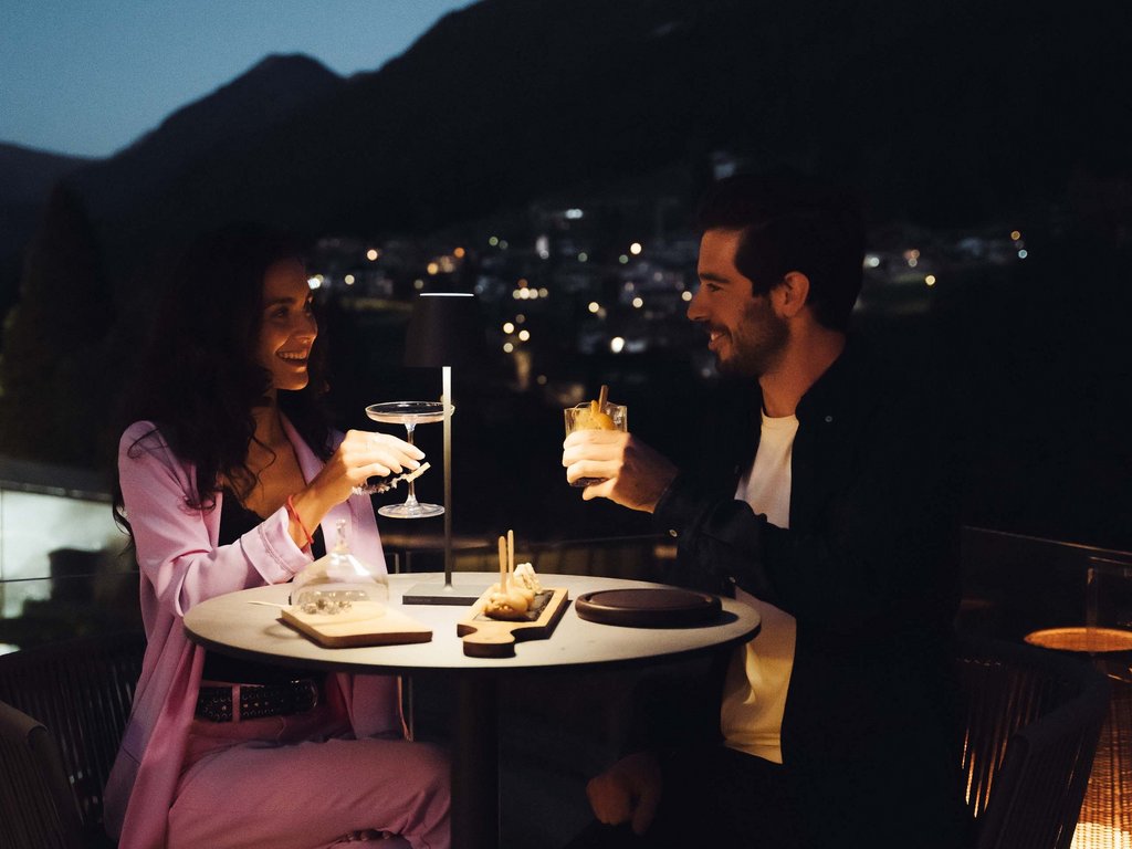 Hotel vicino a Moena: dalle 3 stelle alle 5 stelle! Coppia che brinda con cocktail a cena all'aperto di sera con luci soffuse.