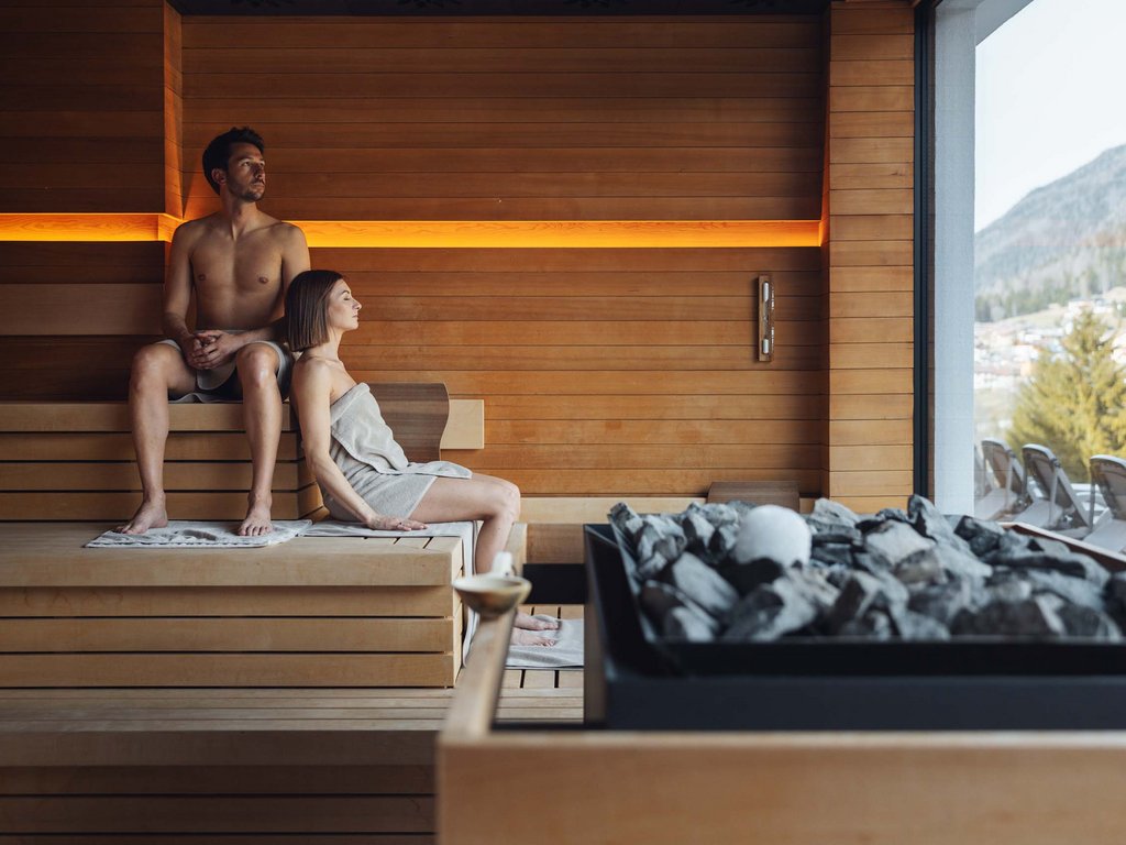 A Soraga il meteo è sempre ottimo per un Day Spa! Coppia rilassata in sauna con vista su montagne e sedie a sdraio