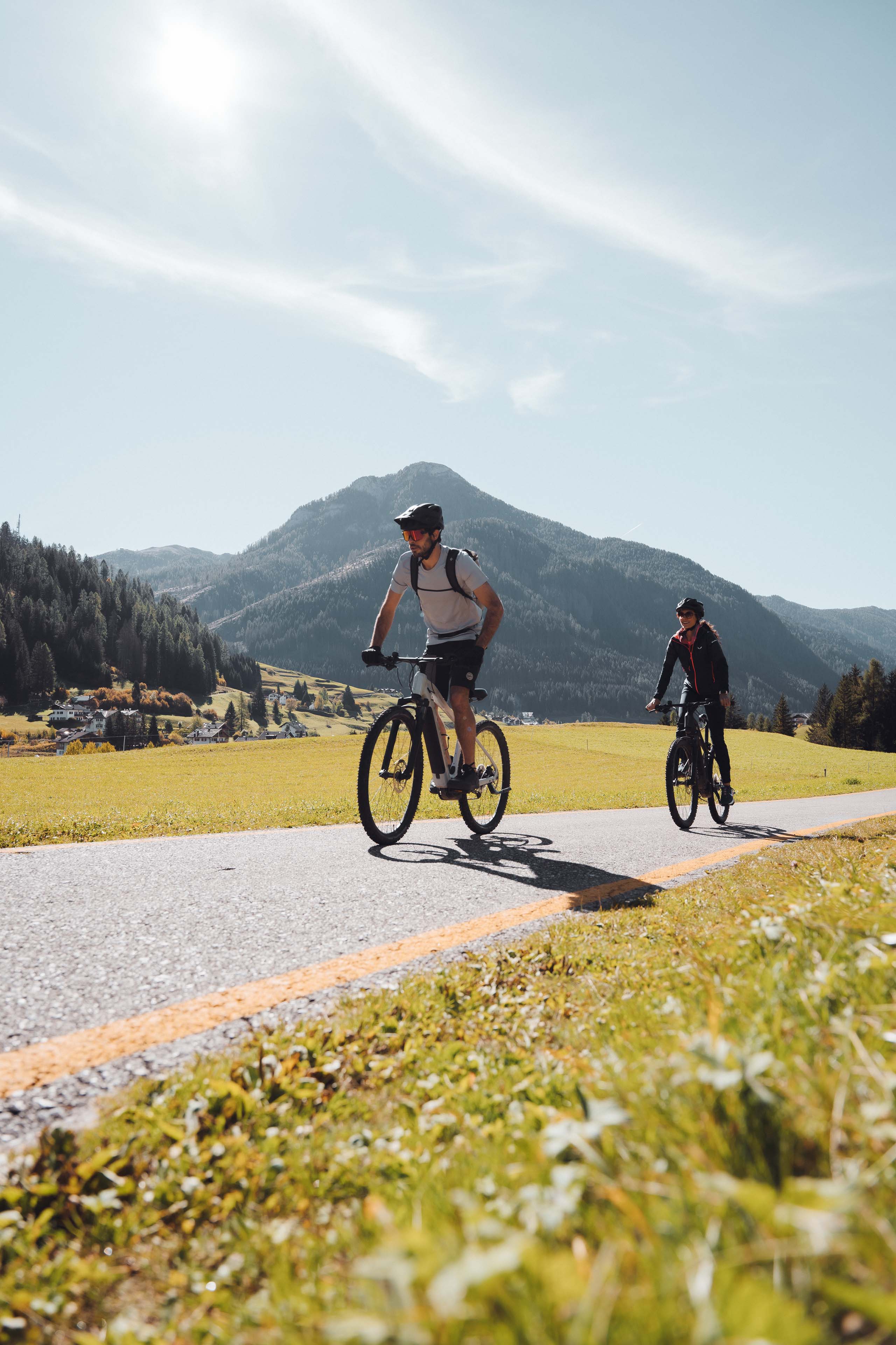 Hotel in Val di Fassa: your Hotel Latemar Two cyclists riding on a scenic road with mountains and fields in the background